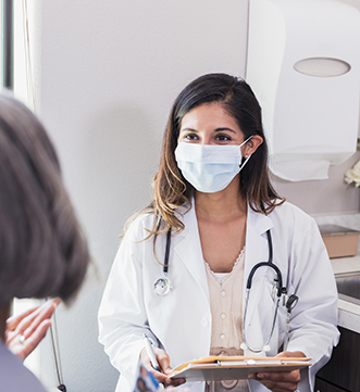 An advanced practice provider wearing a white coat and a surgical mask talking to a patient inside of a medical office.